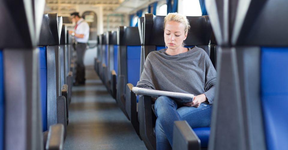 Une femme blonde et détendue est assise seule dans un wagon de train, un journal à la main. Un contrôleur est visible en arrière-plan.